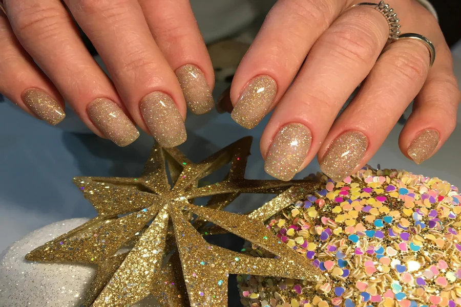 Close-up of holiday glitter nails featuring deep green polish with gold flakes and a holographic accent tip under soft winter lighting