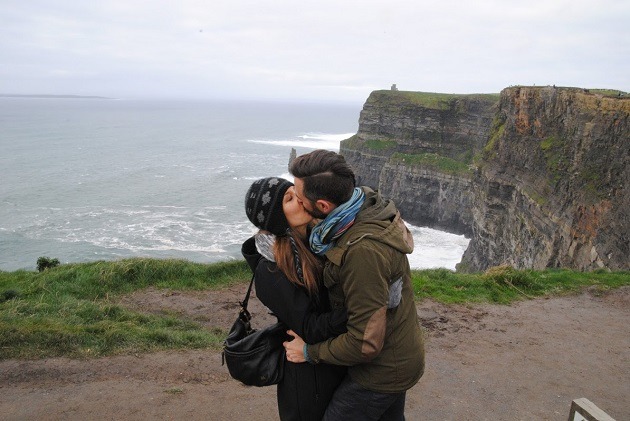 Sunlit cliffs with sheer rock faces dropping to the water below, viewed from a safe overlook with rugged terrain and expansive horizon.