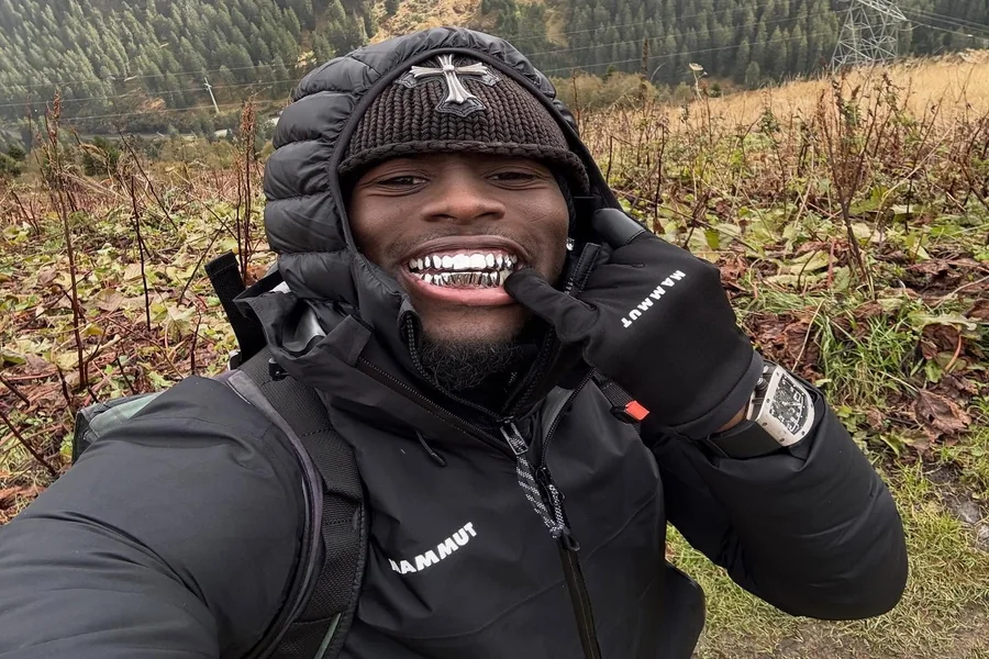 Kai Cenat smiling on a streaming set, standing next to a taller guest with studio lights in the background