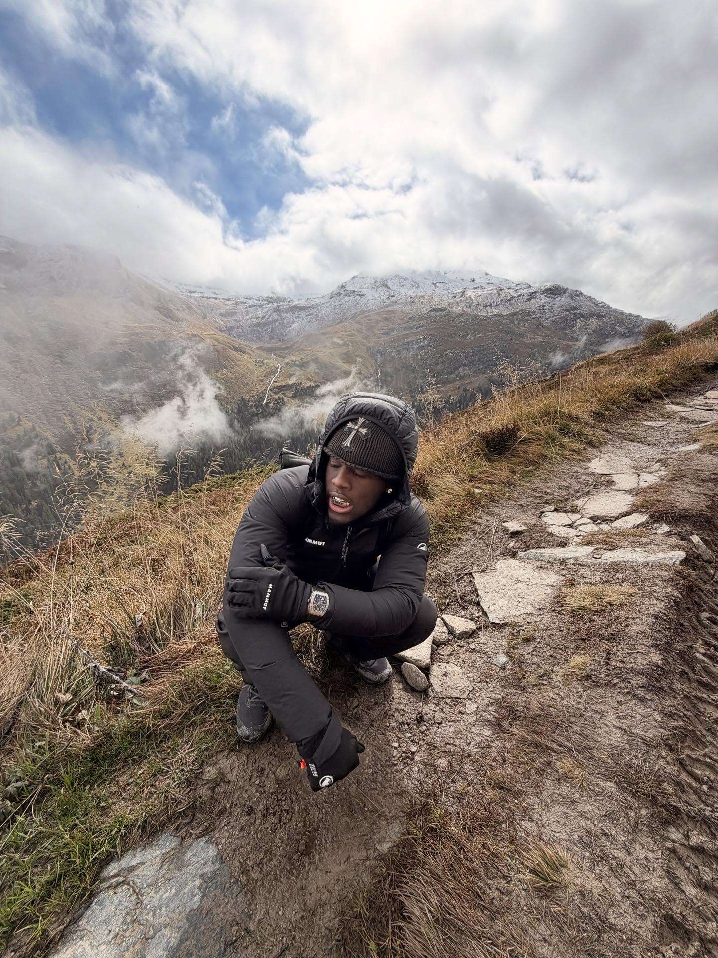 Kai Cenat smiling on a streaming set, standing next to a taller guest with studio lights in the background