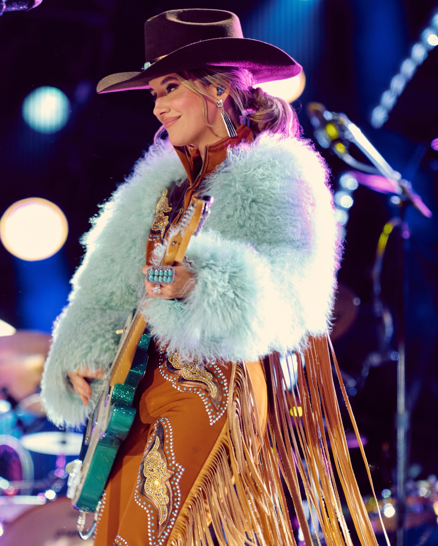 Lainey Wilson performing on stage in vintage-inspired attire with a microphone in hand and warm stage lighting